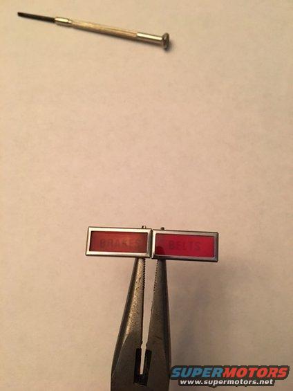 Pliers holding two red warning light lenses labeled BRAKES and BELTS. A small screwdriver is above