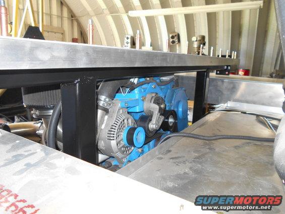 Close-up of a blue engine block with an alternator, belts, and air intake. Some metal sheeting is visible in the foreground