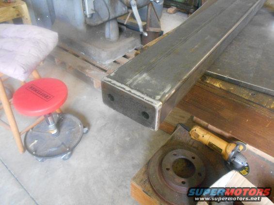 Metal beam on a workbench with a grinder and brake rotor nearby, red stool in background