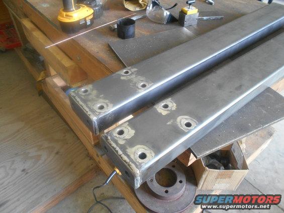 Two metal frame pieces with drilled holes sit on a workbench next to a drill and safety glasses