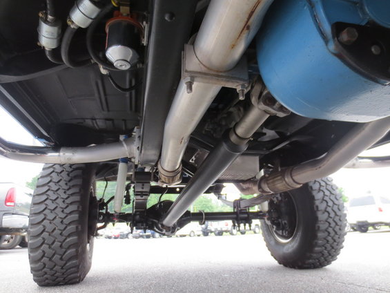 Underside of a lifted truck with large tires, dual exhaust, and a blue fuel tank. Other vehicles are visible in the background