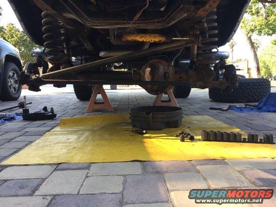 Undercarriage of a vehicle, possibly a truck or SUV, on jack stands with suspension and drivetrain components visible. Tools and a drain pan are on the ground