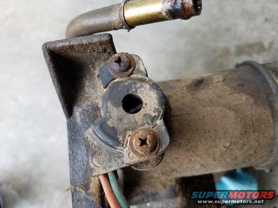 Close-up of a dirty automotive part with a metal hose and wires attached. Two screws hold a black plastic piece in place