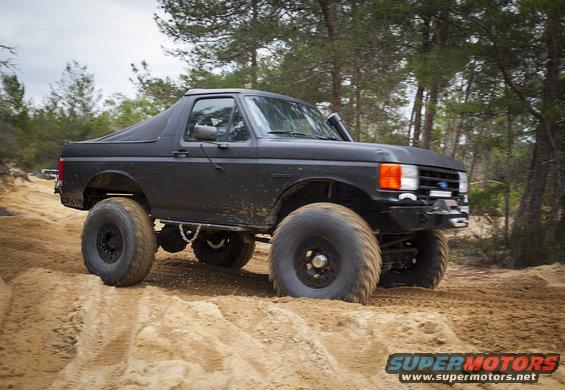 Black Ford Bronco with large tires driving on a sandy trail through trees