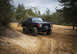 1989 Ford Bronco - Modified black Ford Bronco navigates a sandy off-road trail under an overcast sky.