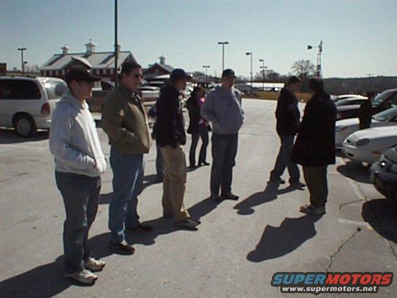 Group of people in a parking lot with a white van and several cars visible