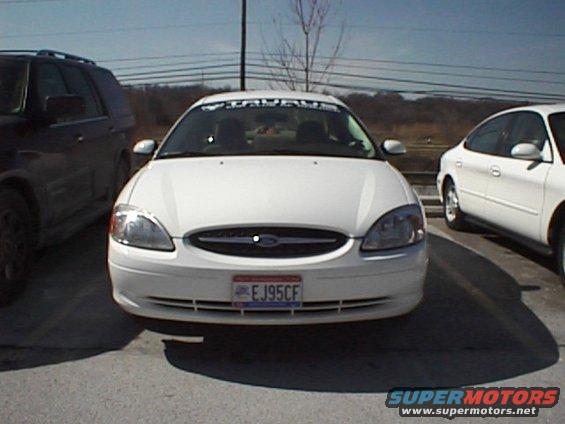 White Ford Taurus with a banner across the windshield, parked next to a dark SUV and another white car