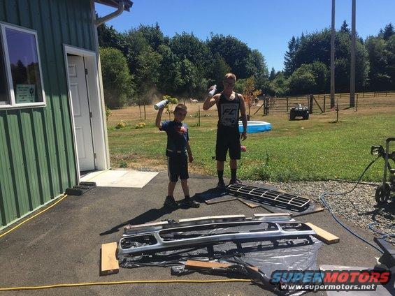 Two boys with spray cans stand near disassembled vehicle grille and bumper parts laid out on the ground