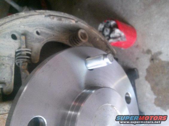 Close-up of a car's brake drum and hub with a wheel stud. A red container with lug nuts is in the background
