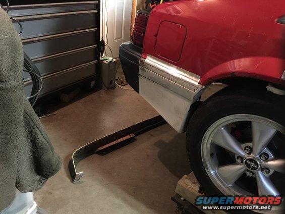 Red car rear quarter panel and bumper area, with a silver bumper strip and a five-spoke wheel. The car is on blocks in a garage