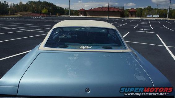 Light blue convertible with a white top, seen from the rear in an empty parking lot. Faded paint and a sticker on the rear window