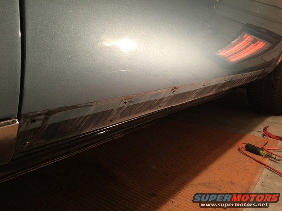 Green car's lower side panel with rust and visible screw holes, showing a partial taillight and tire