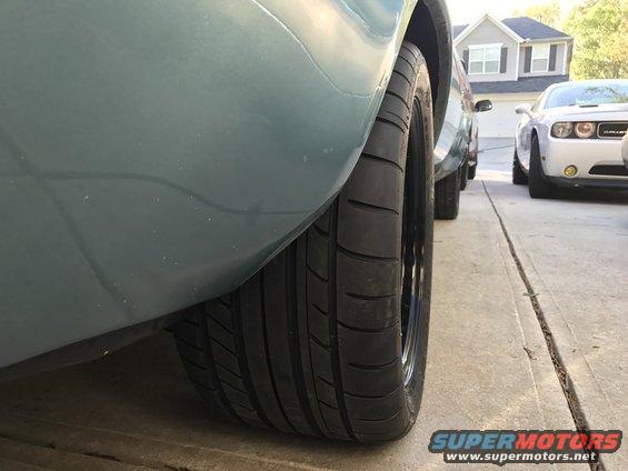 Light blue car's wide rear tire and fender, with a white Dodge Challenger in the background