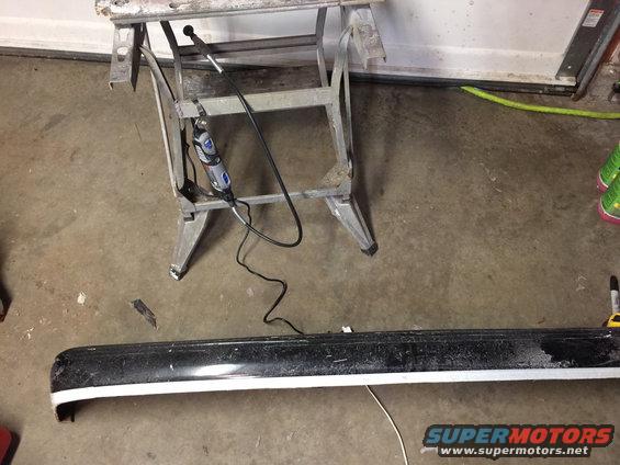 Black car bumper with a white stripe, resting on a concrete floor near a sawhorse holding a Dremel tool