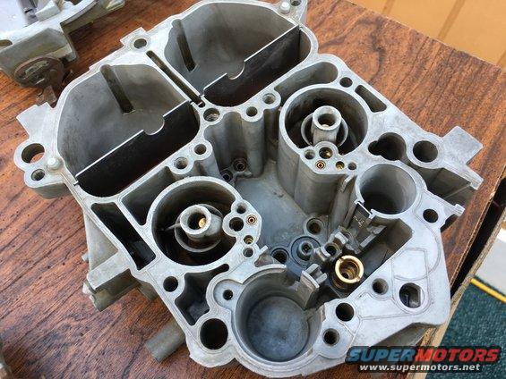 Carburetor part on a wooden surface. A "SUPERMOTORS www.supermotors.net" watermark is visible