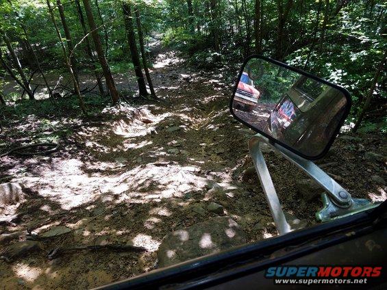 Red truck reflected in side mirror on a muddy, wooded trail