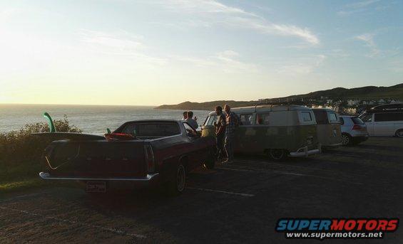 Dark Ford Ranchero parked by the ocean, surfboards in the bed. Other vehicles and people nearby