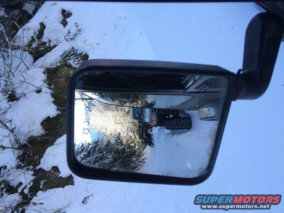 Jeep Wrangler in snow, reflected in side mirror. Large tires, aftermarket bumper visible