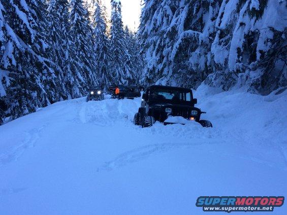 Several Jeeps driving through deep snow on a wooded mountain road. The lead Jeep is a 1995 Jeep YJ