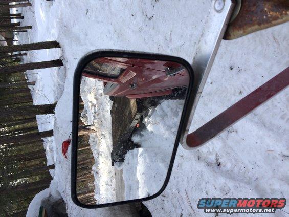 Red vehicle door and interior reflected in side mirror, surrounded by snow and trees