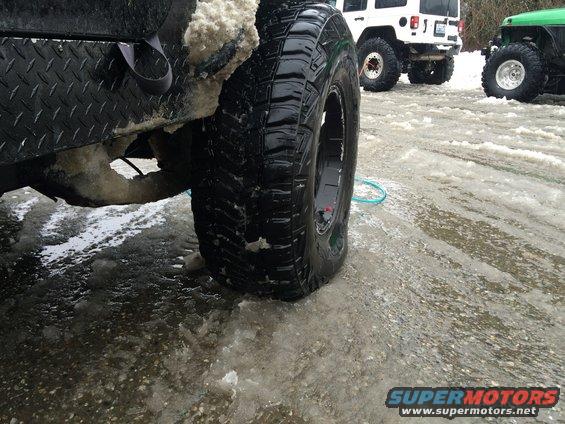 Close-up of a Jeep Wrangler's rear tire and diamond plate bumper in snow. Two other Jeeps are visible in the background
