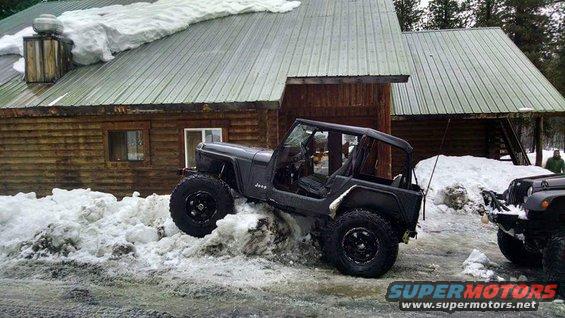 Dark gray Jeep Wrangler YJ with large tires stuck in snowdrift in front of log cabin