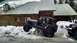 1995 Jeep YJ - A dark gray Jeep Wrangler YJ navigates a snow-covered area near a log cabin, with another Jeep visible in the background.