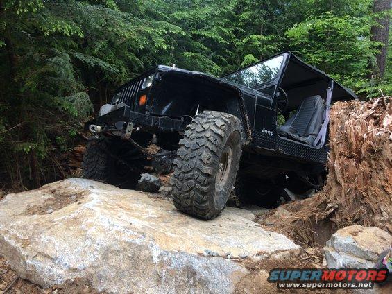 Black Jeep Wrangler navigating large rocks on a wooded trail. The vehicle has large off-road tires and appears to be climbing over an obstacle