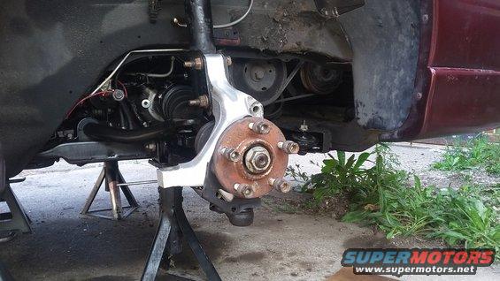 Car's front wheel hub and suspension assembly on jack stands, showing brake rotor, CV axle, and engine belt