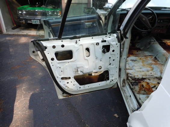 Interior of a car with door removed, showing exposed door frame and rusted floor. Another green car is visible in the background