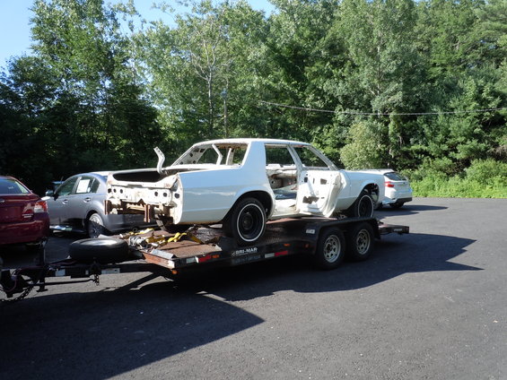 White sedan chassis on a trailer, doors open, stripped interior, black wheels, parked outdoors