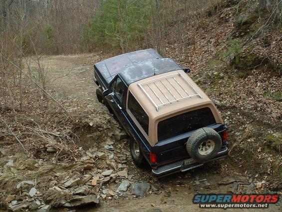 Dark blue 1987 Ford Bronco with tan top and roof rack driving on a rocky, wooded trail