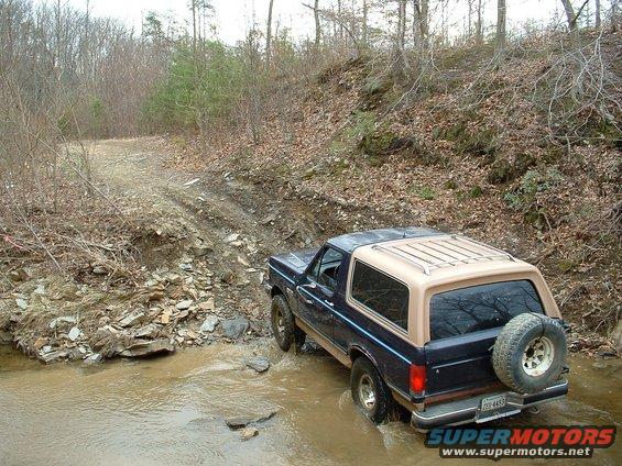 1987 Ford Bronco driving through a shallow stream on a wooded trail. Spare tire mounted on rear door