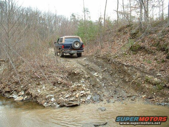 Blue and tan Ford Bronco driving up a muddy, rocky trail next to a stream