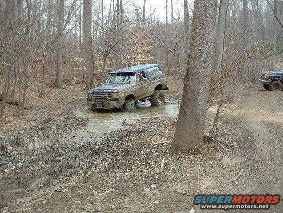 Mud-covered 1987 Ford Bronco driving through a muddy trail in a forest, with another vehicle visible in the background