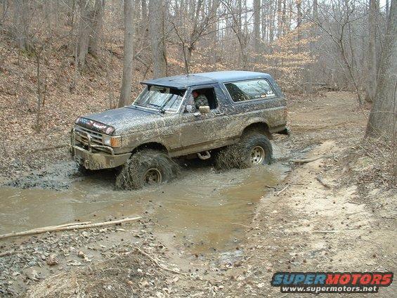 Muddy blue 1987 Ford Bronco driving through a water-filled ditch