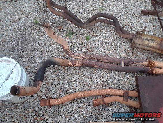 Rusty exhaust pipes and muffler laid out on gravel. A white bucket is to the left