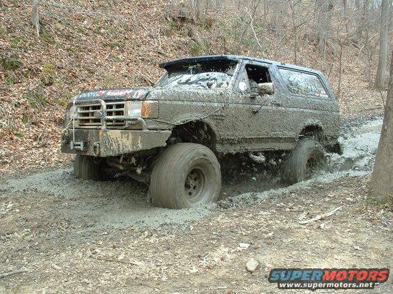Mud-covered 1987 Ford Bronco with large tires navigating a muddy off-road trail