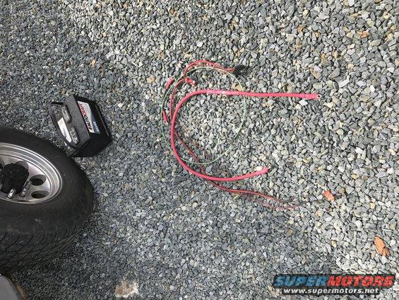 Battery and loose red and green cables on gravel next to a tire and wheel
