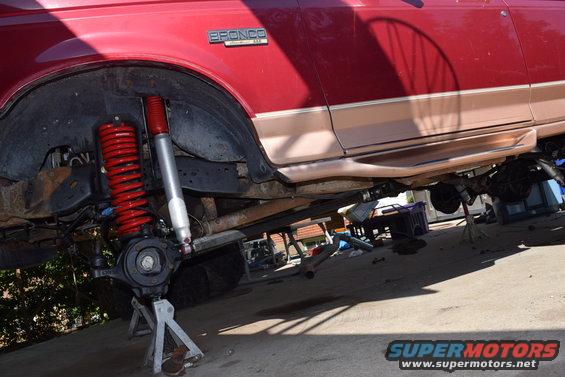 Red Ford Bronco with tan trim, front suspension removed and supported by jack stands, red coilover shocks visible