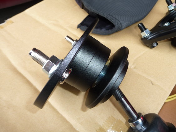 Close-up of black automotive suspension components with threaded bolts and nuts. A grey fabric bag is in the background