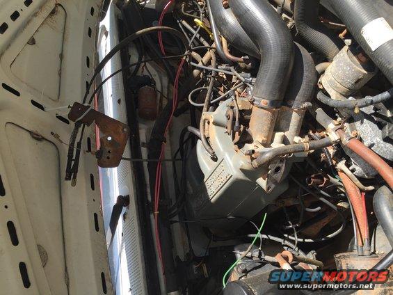Engine bay with hoses, wires, and engine components visible. Some text on a gray component