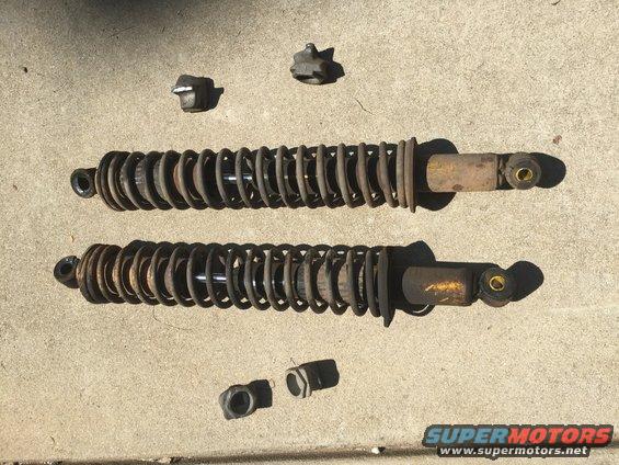 Two rusty shock absorbers with yellow bodies and coiled springs, along with four mounting hardware pieces, lie on a concrete surface