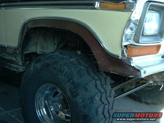 Close-up of a lifted Ford Bronco's front fender, showing a large off-road tire and chrome wheel. The paint is two-tone, beige and brown