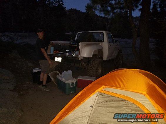 White pickup truck parked at a campsite with camping gear and a tent in the foreground. A person stands by the truck bed