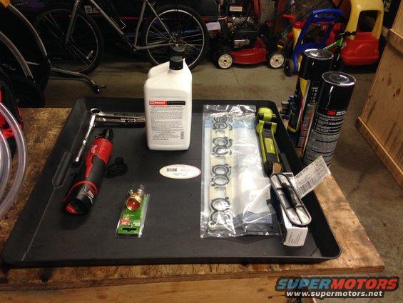 Black tray with automotive parts: gasket set, fluid bottle, wrench, power tool, and spray cans