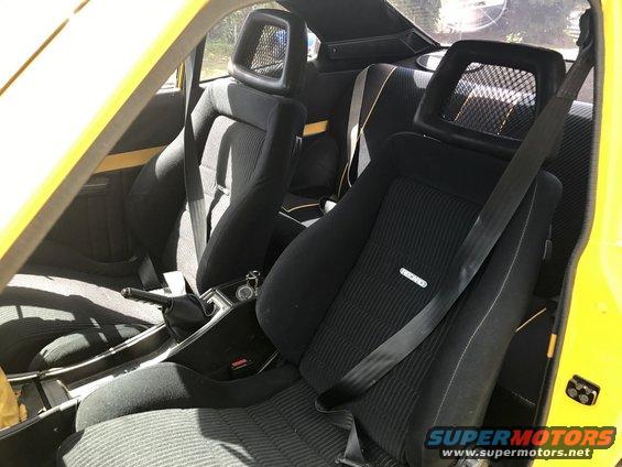 Yellow car's interior showing black seats with headrests and seatbelts. Recaro logo visible on passenger seat