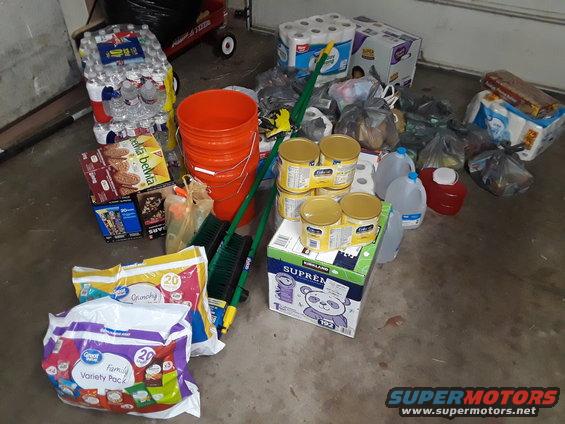 Collection of water bottles, toilet paper, canned goods, cleaning supplies, and packaged food items on a concrete floor