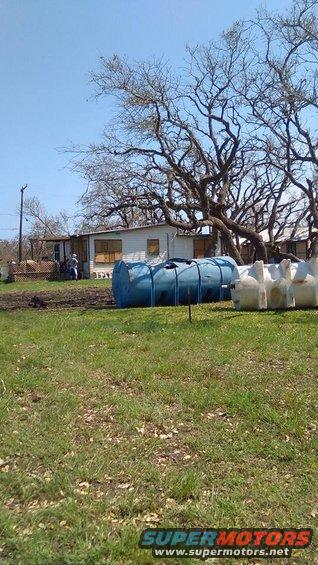 Blue and white water tanks sit in front of a house with large trees in the background