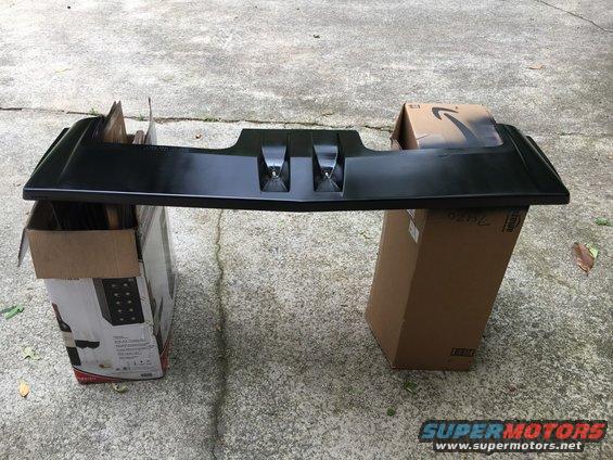 Black plastic car part, possibly a bumper or spoiler, resting on cardboard boxes